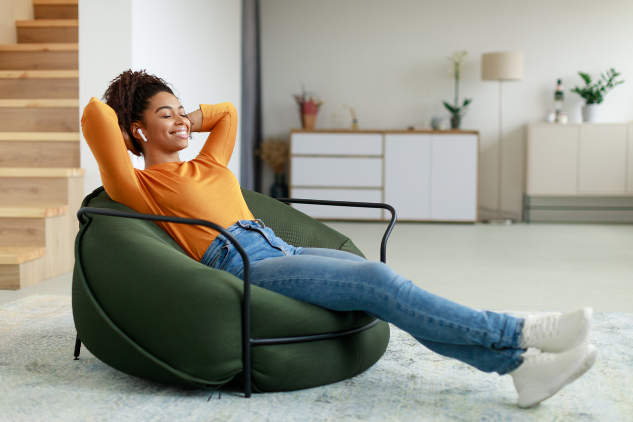Rest And Relax Concept. Calm black woman sitting on bean bag, listening to music, audio book with wireless earphones,